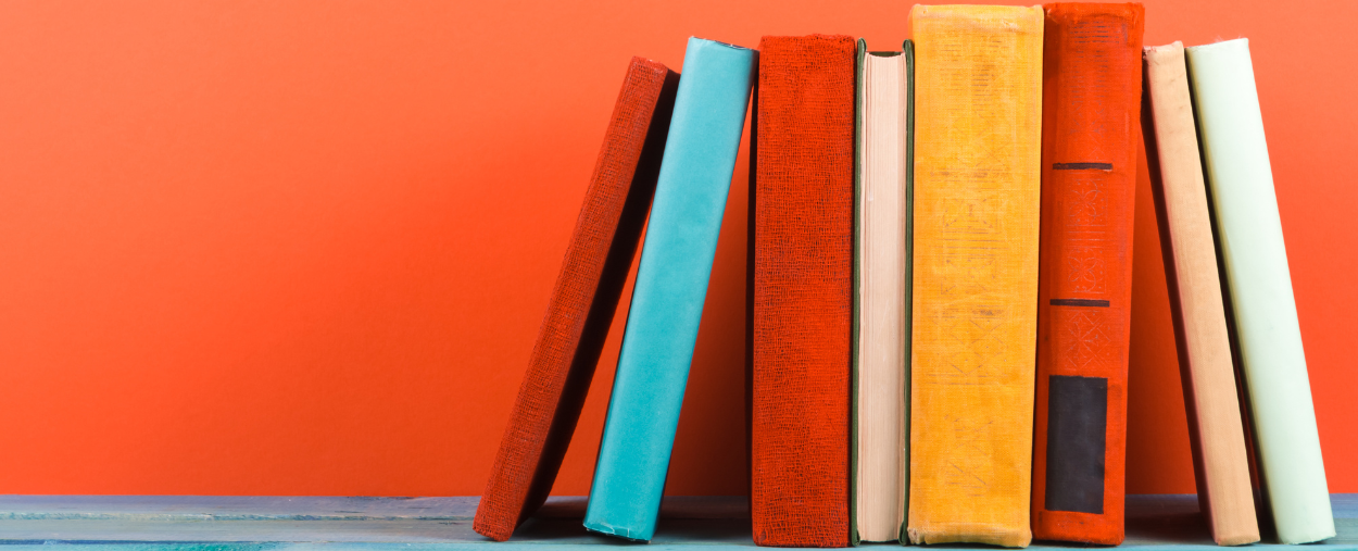 a stock photo of 8 books propped up. The books are of various size and colors such as red, blue, orange, yellow, and white. The books are set against an orange background