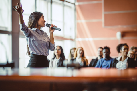 a stock photo of a lady giving a speech in front of a group of people. The lady has long dark brown hair and is wearing a blue button down shirt. She is holding a microphone and pointing with her right hand. The group is in the background and unfocused.
